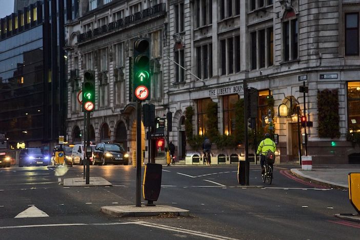 London street scene at dusk in Tower Hill showing traffic signals displaying green lights with a cyclist in high-visibility clothing riding through an intersection. The Liberty Bounds JD Wetherspoon pub is visible in the background alongside historic buildings, with multiple vehicles on the road and street lighting illuminating the urban environment.