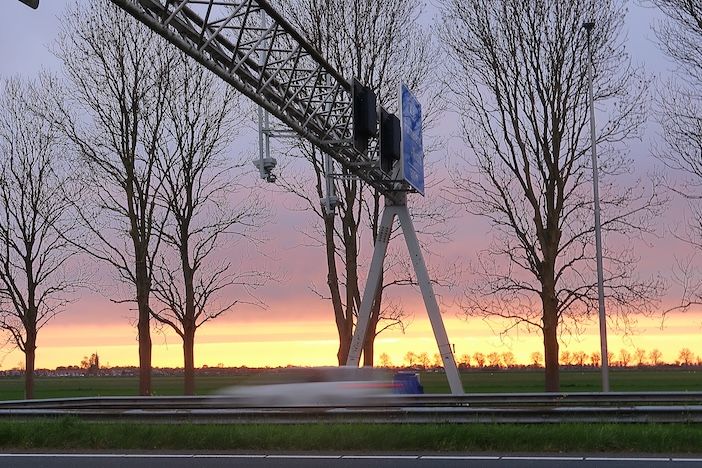 Close-up of white average speed enforcement camera mounted on highway gantry with infrared illuminators, vehicle with headlights passing underneath at dusk