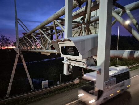 Highway gantry structure with mounted enforcement equipment at sunset, silhouetted against orange and pink sky with bare winter trees alongside Dutch motorway