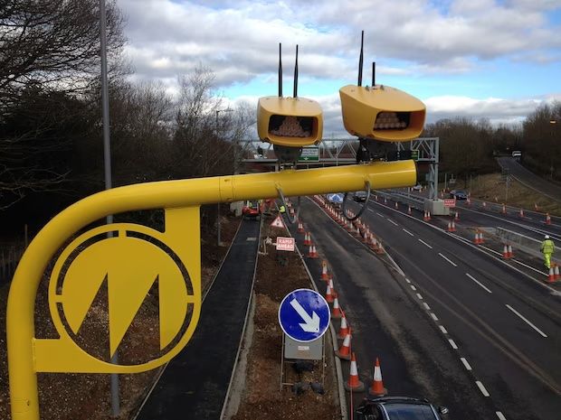  Jenoptik average speed enforcement camera system installation on motorway showing distinctive yellow gantry mounted across carriageway with dual camera units in yellow housings monitoring traffic lanes with roadworks and traffic cones visible below