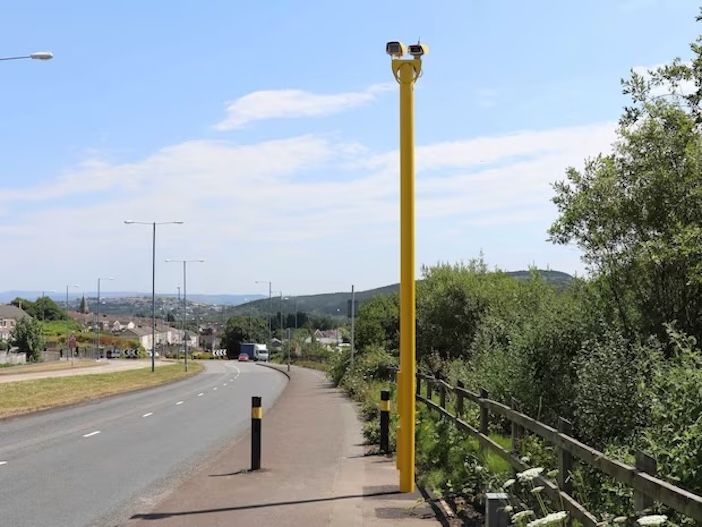 Jenoptik speed enforcement camera mounted on tall yellow pole beside residential road and pedestrian footpath, showing dual camera heads at height against clear sky with housing estate and hills visible in background