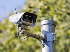 Jenoptik roadside speed enforcement camera mounted on galvanised steel pole showing white weatherproof housing with black front panel, infrared LED array and external antenna for wireless connectivity against blurred foliage background