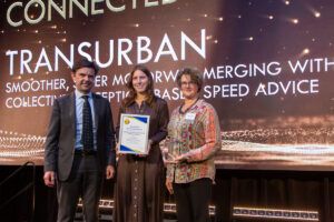 Three people standing on stage at the ITS Australia 16th Annual Awards ceremony, with the centre person holding a framed certificate for Transurban's Connected and Automated Vehicle Award. A presentation screen behind displays the Transurban name and project details.