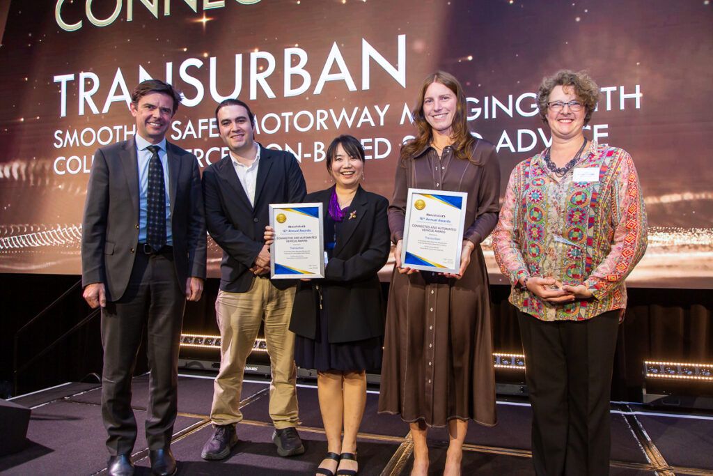 Five people standing on stage at the ITS Australia Awards, two holding framed certificates for Transurban's Connected and Automated Vehicle Award, with the award category and project details displayed on the screen behind them