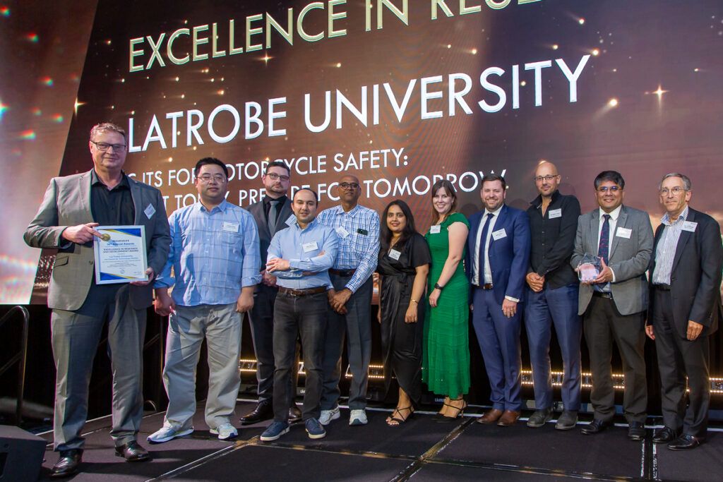 A large group of approximately eleven people standing on stage at the ITS Australia Awards, with one person holding a framed certificate. The screen behind reads "Excellence in Research" and displays La Trobe University's name and its C-ITS for motorcycle safety project.