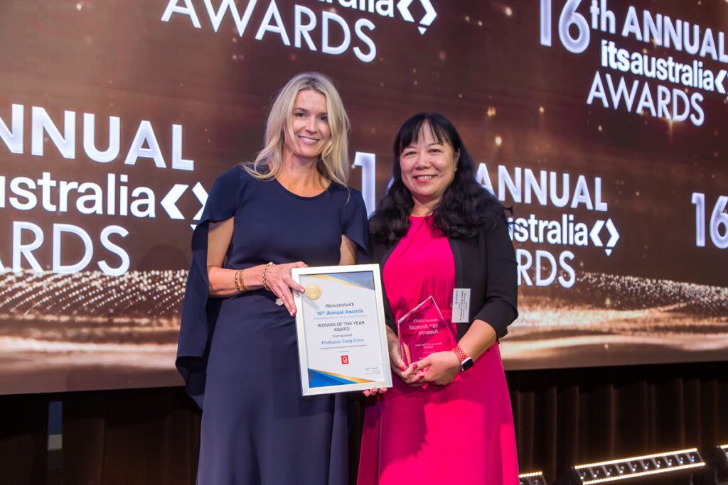 : Two women standing on stage at the ITS Australia 16th Annual Awards. One holds a framed certificate reading "Woman of the Year Award – Distinguished Professor Fang Chen" and the other holds a glass trophy.
