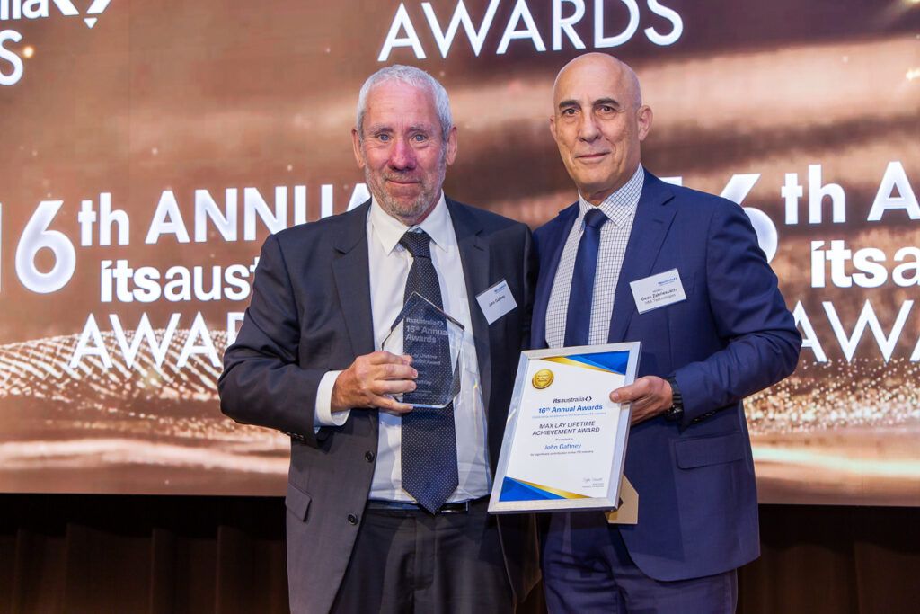 Two men standing on stage at the ITS Australia 16th Annual Awards, one holding a glass trophy and the other holding a framed certificate reading "Max Lay Lifetime Achievement Award – John Gaffney".