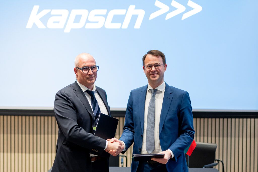 Two men in business suits shake hands in front of a Kapsch corporate backdrop. The man on the left wears a dark suit and round glasses, whilst the man on the right wears a blue suit. Both hold dark folders and are smiling at the camera in what appears to be a formal contract signing ceremony.