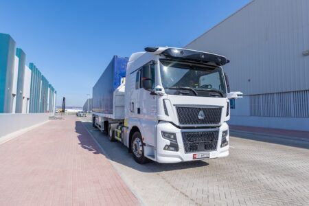 Abu Dhabi launches first autonomous truck pilot in MENA logistics sector A white autonomous heavy goods truck with sensor equipment mounted on the roof cab, pulling a flatbed trailer loaded with a shipping container, parked on a brick-paved road outside a large industrial warehouse facility under a clear blue sky. An Abu Dhabi licence plate is visible on the front of the vehicle.