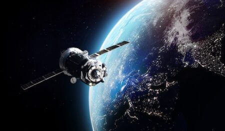 Computer-generated illustration of a telecommunications satellite with extended solar panels orbiting Earth at night, with city lights visible on the planet's surface below and the curve of the atmosphere glowing blue.