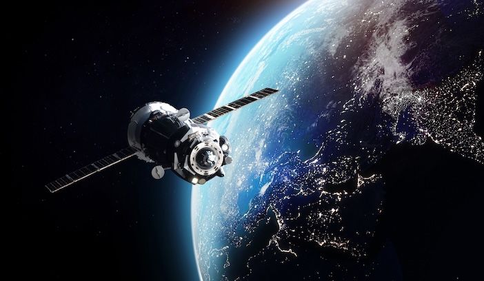 Computer-generated illustration of a telecommunications satellite with extended solar panels orbiting Earth at night, with city lights visible on the planet's surface below and the curve of the atmosphere glowing blue.