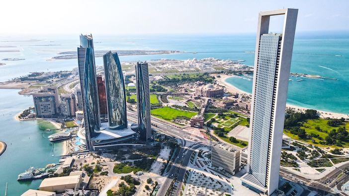 Aerial view of Abu Dhabi's city centre skyline, showing a cluster of modern high-rise towers and the Emirates Palace hotel set among green landscaped grounds, with the Arabian Gulf coastline and islands visible in the background under a hazy blue sky