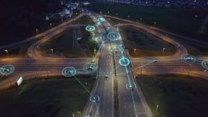 An aerial night-time view of a multi-lane road interchange with vehicles visible, overlaid with blue circular connectivity icons and lines linking vehicles across the network, illustrating vehicle-to-infrastructure communication.