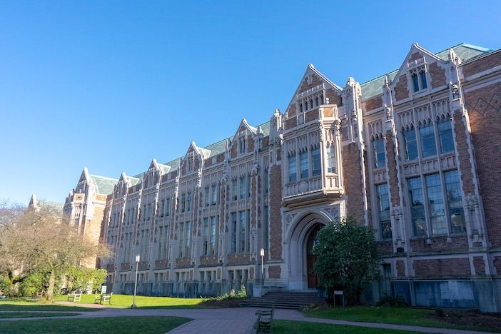Exterior view of a Gothic Revival stone building on the University of Washington campus in Seattle, with ornate arched windows, decorative stonework, and a green lawn in the foreground under a clear blue sky.