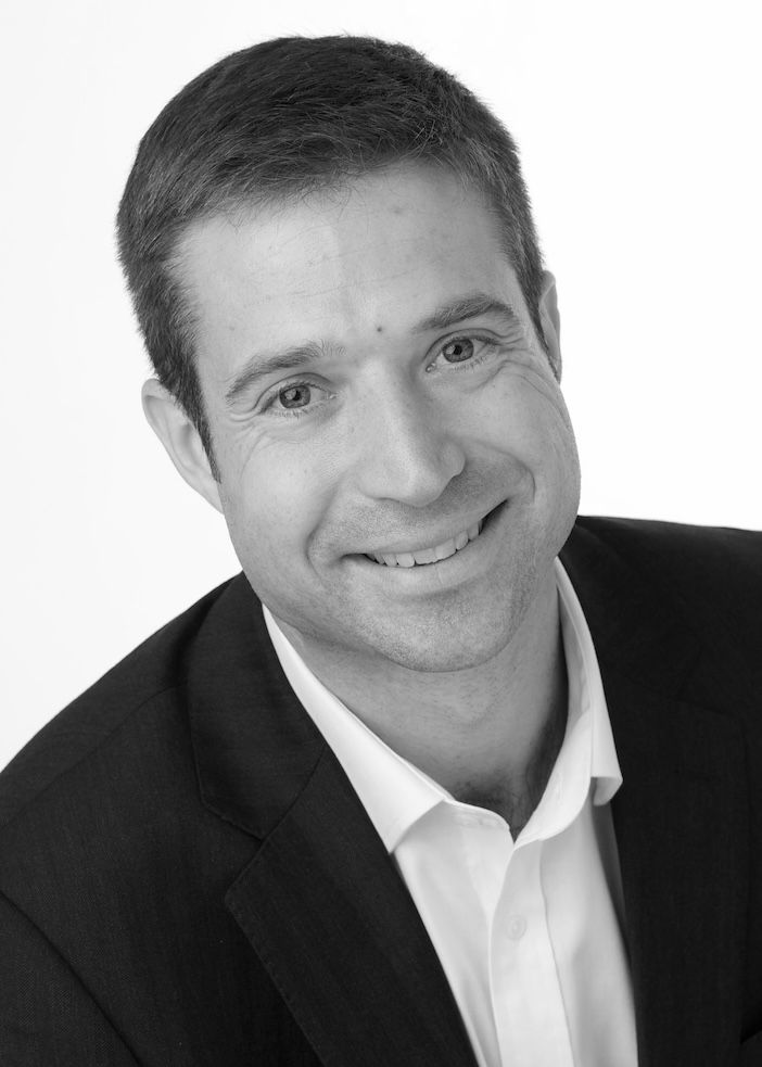 Black and white headshot of Andy Hutt, CEO of Triopsis, smiling at the camera wearing a dark blazer and open-collar white shirt.