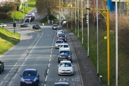 An aerial view of a multi-lane road in Plymouth showing a queue of vehicles travelling in both directions. Overhead gantries with road safety cameras are visible on the right-hand side of the carriageway, with green verges and trees lining the route.