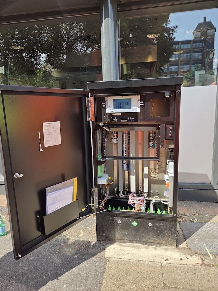 Open roadside traffic controller cabinet showing internal electronic components including a display screen, circuit boards, wiring looms, and modular equipment racks. SWARCO PTC-3 documentation is visible on the inside of the cabinet door. The cabinet is mounted on a concrete plinth at a street location with glass building frontage visible in the background.