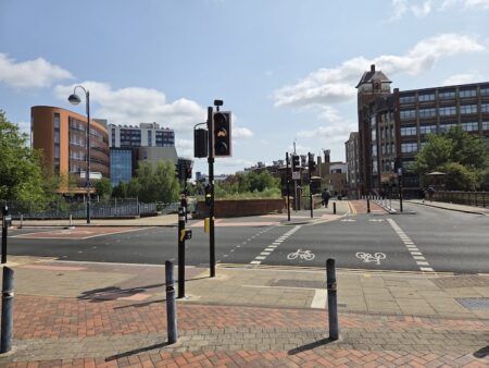 Urban junction with dedicated cycle lanes and traffic signals in a UK city centre. A bicycle traffic light displays an amber cycle symbol in the foreground. The road features painted cycle lane markings and is bordered by a mix of modern curved orange-clad buildings and traditional red brick Victorian architecture. Pedestrian bollards separate the pavement from the carriageway.