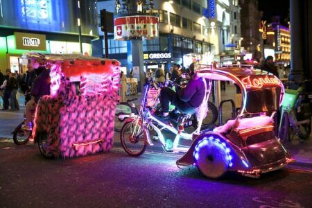 London introduces first pedicab regulations with fare caps and noise ban Two brightly lit and decorated pedicabs parked on a London street at night, surrounded by illuminated shop fronts including an M&M's store and a Greggs. The vehicles feature colourful LED lighting and decorative fur trim, with a driver visible on one of the pedicabs.