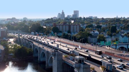 Aerial photograph of Washington Bridge in Providence, Rhode Island, showing the historic arched stone bridge carrying multi-lane traffic across a river, with the city skyline visible in the background.