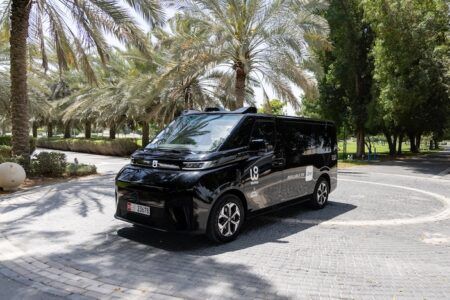 A black autonomous taxi vehicle with white number 19 livery and WeRide branding parked on a paved area surrounded by palm trees and greenery in Abu Dhabi. The compact vehicle features sensors and cameras mounted on its roof for autonomous navigation, with Abu Dhabi license plates visible.