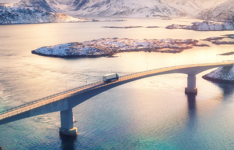 Aerial view of a truck crossing a curved bridge over turquoise water in a remote Arctic landscape, with snow-covered mountains and small islands visible in the background during winter sunrise.