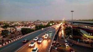 Aerial view of a multi-lane urban expressway in India at dusk, with vehicles travelling in both directions under street lighting, elevated transit infrastructure visible to the right and low-rise city buildings stretching to the horizon.