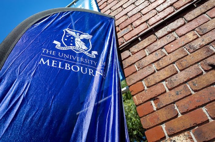 A blue branded banner bearing the University of Melbourne crest and name, mounted against a red brick wall under a clear blue sky.