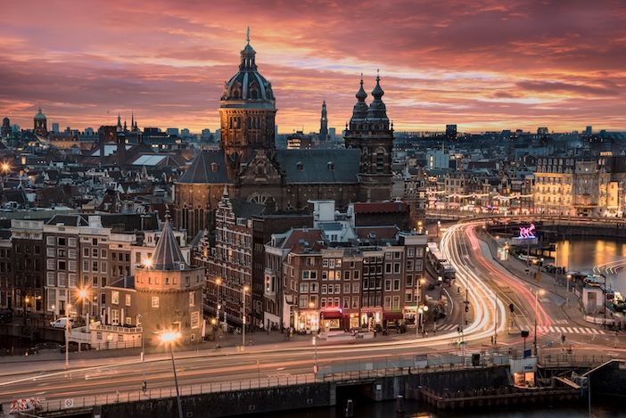 Aerial view of Amsterdam at dusk, showing the Basilica of Saint Nicholas and surrounding historic city centre, with light trails from traffic on the road below and a dramatic red and orange sky above.