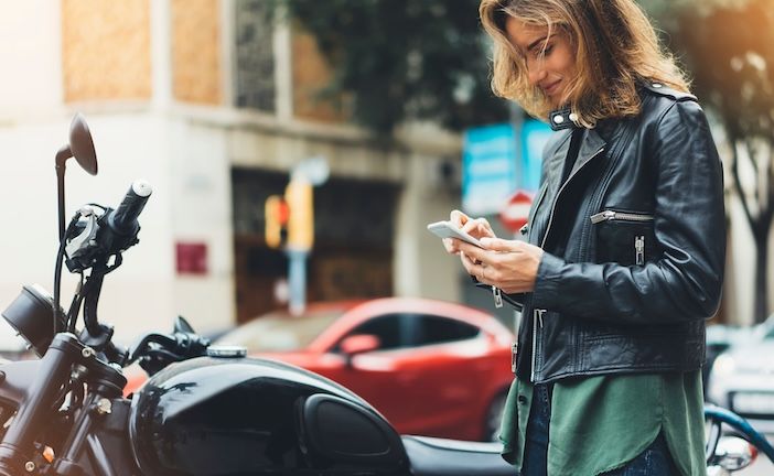 A woman in a leather jacket stands beside a motorcycle on an urban street, looking at her smartphone.