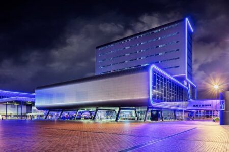 Exterior of a large modern exhibition centre at night, illuminated with blue neon lighting along its facade and roofline, with a wide paved forecourt in the foreground.