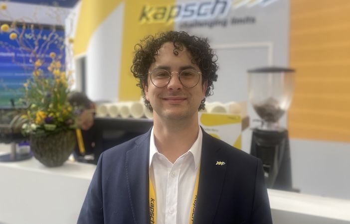A young man with curly dark hair and glasses, wearing a navy blazer and white shirt, smiles at the camera in front of the Kapsch TrafficCom exhibition stand at Intertraffic Amsterdam 2026.