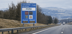 A blue motorway services sign on the M6 showing the Moto Lancaster Services at half a mile, displaying logos for Costa, M&S, Greggs and Burger King, along with current petrol and diesel fuel prices, positioned beside a rural dual carriageway with hills in the background.