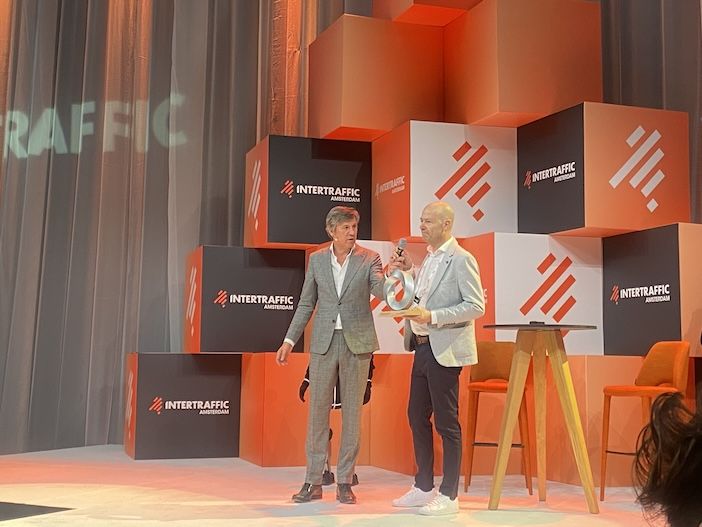 Two men in suits stand on stage at Intertraffic Amsterdam in front of an orange and white branded cube backdrop; one holds a microphone and a circular silver trophy.