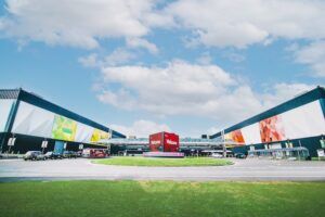Exterior view of the National Exhibition Centre (NEC) in Birmingham, showing two large parallel exhibition halls with colourful geometric murals, a central roundabout with a red welcome cube, coaches and taxis in the foreground, and a blue sky with scattered clouds above.
