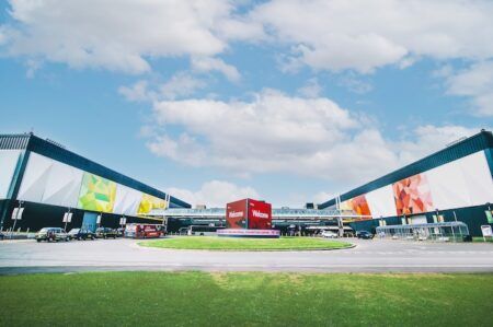 ITS World Congress and Highways UK to co-locate at NEC Birmingham in 2027 Exterior view of the National Exhibition Centre (NEC) in Birmingham, showing two large parallel exhibition halls with colourful geometric murals, a central roundabout with a red welcome cube, coaches and taxis in the foreground, and a blue sky with scattered clouds above.
