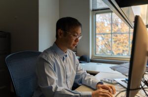A man wearing glasses and a light blue shirt sits at a desktop computer in an office, working at a keyboard. Autumn foliage is visible through a window behind him