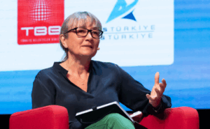 A woman with grey hair and glasses sits in a red chair on a conference stage, gesturing with one hand while holding a notebook. Sponsor logos including TBB and ITS Türkiye are visible on a screen behind her.