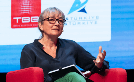 A woman with grey hair and glasses sits in a red chair on a conference stage, gesturing with one hand while holding a notebook. Sponsor logos including TBB and ITS Türkiye are visible on a screen behind her.