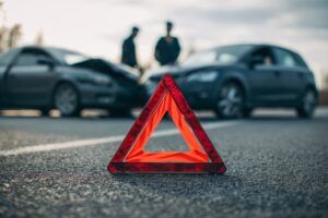 Two individuals converse beside a checkpoint warning device, with wrecked cars behind them amid overcast skies, capturing the somber mood of a recent collision.