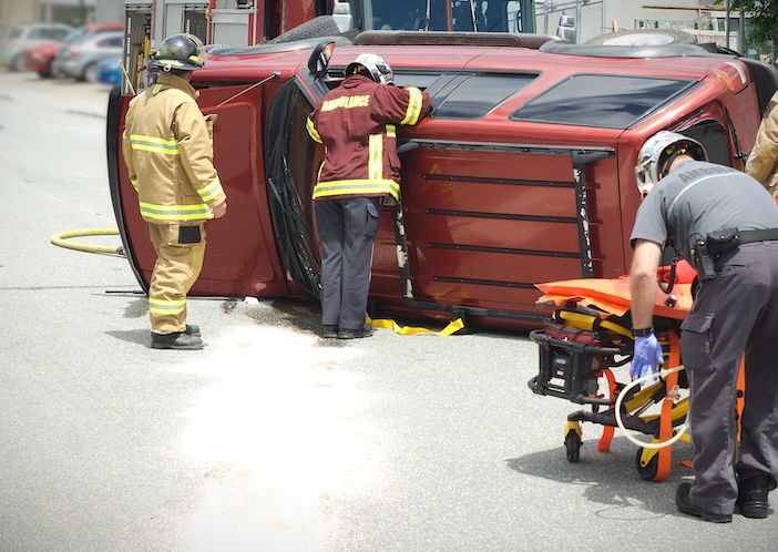 Emergency responders attending an overturned red vehicle on a road, with a firefighter, ambulance crew member, and police officer visible alongside a stretcher