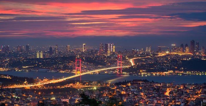 Night panorama of Istanbul showing the illuminated 15 July Martyrs Bridge spanning the Bosphorus Strait, with the city's European and Asian skylines visible on either side under a vivid red and purple sky.