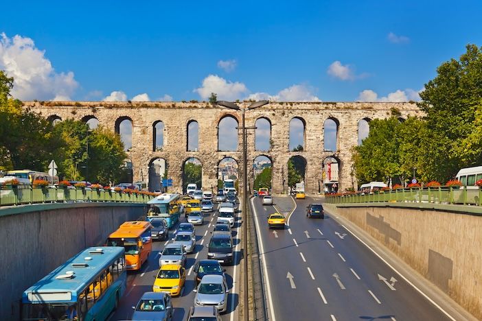 Heavy traffic on a multi-lane road passing beneath the ancient Valens Aqueduct in Istanbul, with buses, taxis and cars queueing in both directions under a blue sky