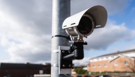 A white ANPR camera mounted on a metal pole against a partly cloudy sky, with a brick building visible in the background