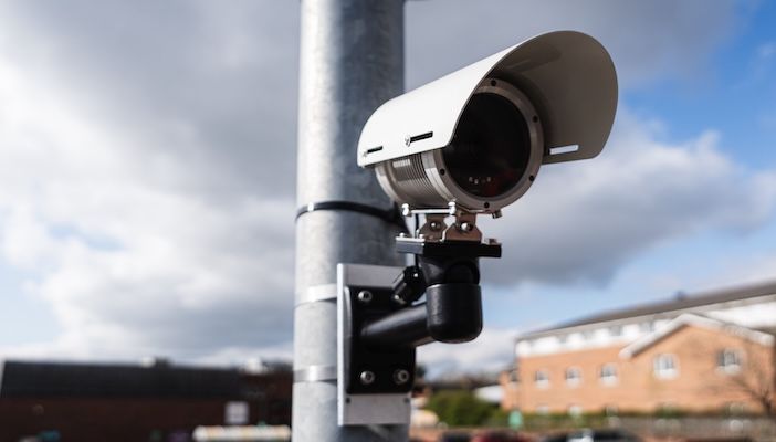 A white ANPR camera mounted on a metal pole against a partly cloudy sky, with a brick building visible in the background