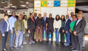 A group of approximately 18 people, including Ghanaian transport officials and UK representatives, pose for a photograph inside TRL's Woolwich Living Lab. Behind them is a branded display board featuring the logos of Cisco, Loughborough University, dg:cities, Transport for London, and Cubic.