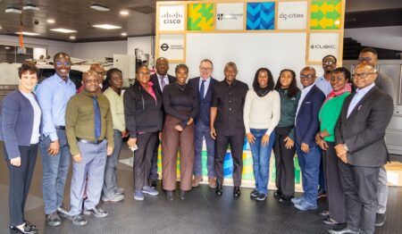 A group of approximately 18 people, including Ghanaian transport officials and UK representatives, pose for a photograph inside TRL's Woolwich Living Lab. Behind them is a branded display board featuring the logos of Cisco, Loughborough University, dg:cities, Transport for London, and Cubic.