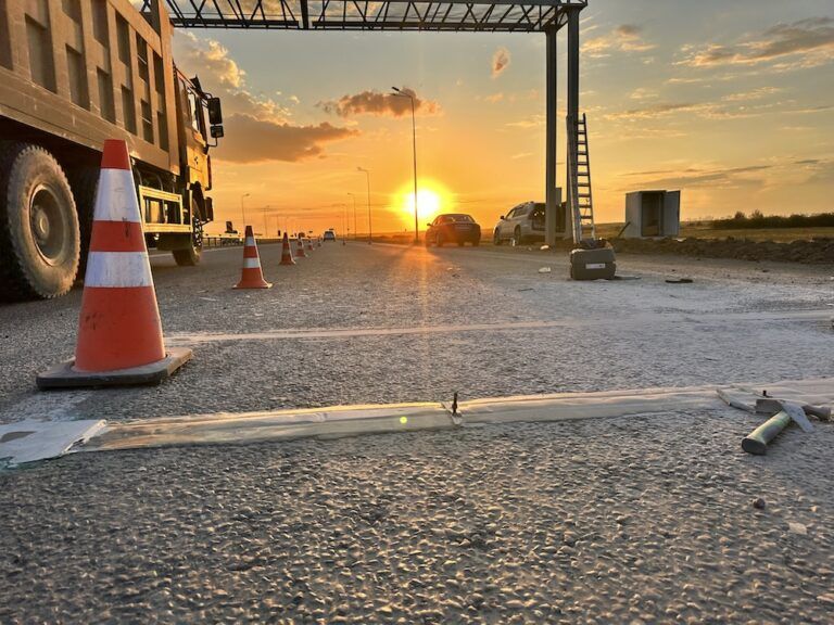 Strain gauge strip sensor laid into the road surface at a Kazakhstan HS-WIM site at sunset, with traffic cones, a parked dump truck and an enforcement gantry in the background
