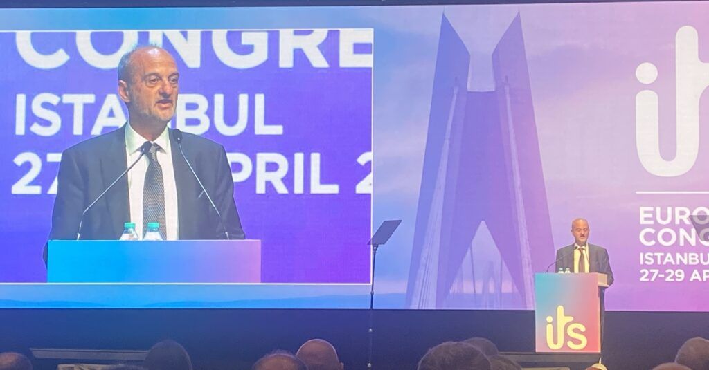 A speaker addresses delegates from a podium at the ITS European Congress in Istanbul, with the congress branding and city logo visible on a large screen behind him, and a second speaker visible at a podium in the background to the right