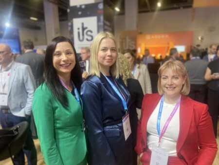Three women stand together smiling at a busy exhibition hall at the ITS European Congress in Istanbul, with an ITS banner visible in the background.